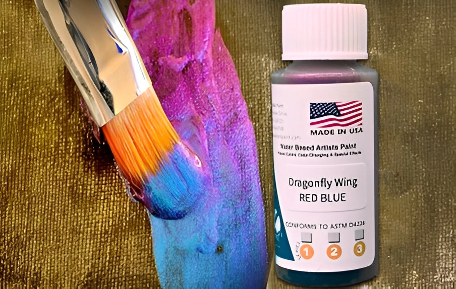 A paintbrush with bright blue and pink paint next to a bottle of "Dragonfly Wing Red Blue" water-based artist paint, labeled with an American flag and "Made in USA.