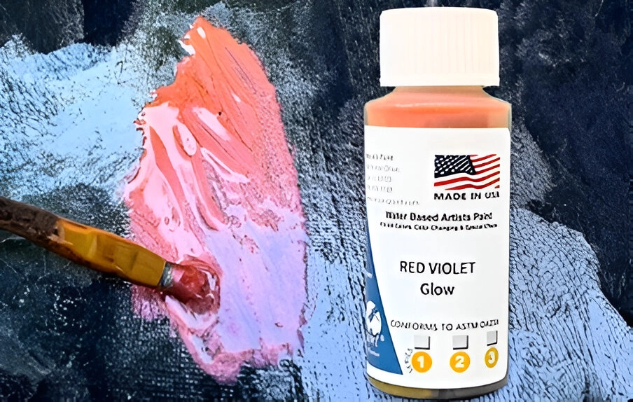 A bottle labeled "Red Violet Glow Water Based Artists Paint" with an American flag, next to a paintbrush and a small smear of pinkish paint.