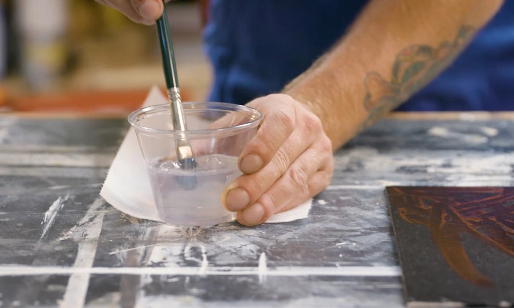 A person with tattoos is rinsing a paintbrush in a plastic cup filled with water on a white paper towel, next to an artistic canvas.