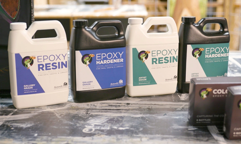 Four containers of epoxy resin and hardener, labeled “Gallery Grade” and “Artist Grade”, placed on a workshop table with other crafting supplies in the background.