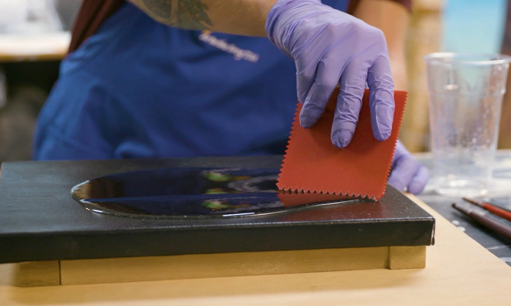 A person wearing gloves and an apron uses a red, serrated-edge tool to spread a liquid across a black surface on a wooden stand. Two paintbrushes and a clear plastic cup are in the background.