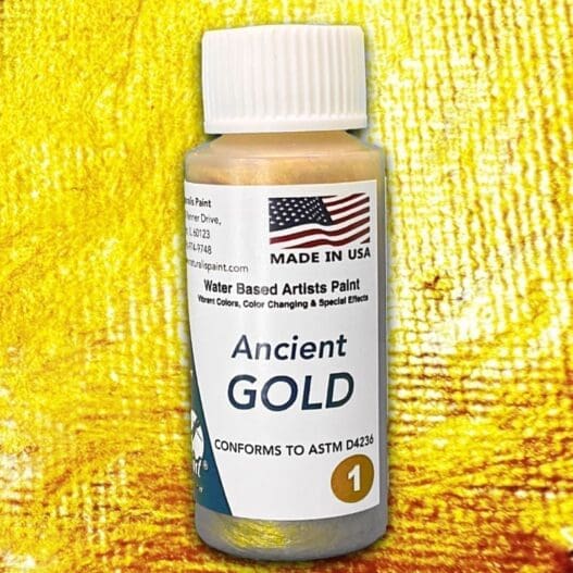 A small bottle of "Ancient Gold" water-based artist paint, featuring a U.S. flag and conforming to ASTM D4236 standards, set against a yellow textured background.