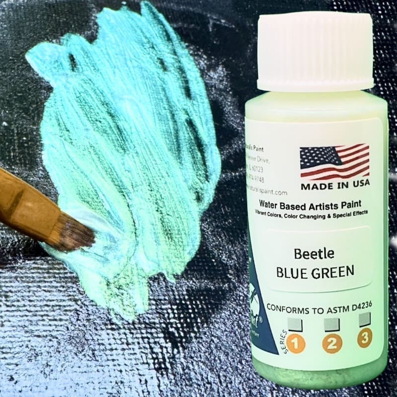 A bottle of Bettle Blue Green water-based artist paint stands next to a brush applying the vibrant color on a black canvas. The label on the bottle indicates it conforms to ASTM D4236 and is made in the USA.