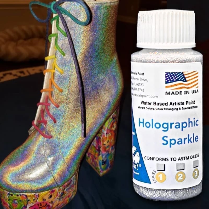 A boot with holographic rainbow accents is displayed next to a bottle of "Holographic Sparkle" water-based paint, featuring an American flag and a label stating it is made in the USA.