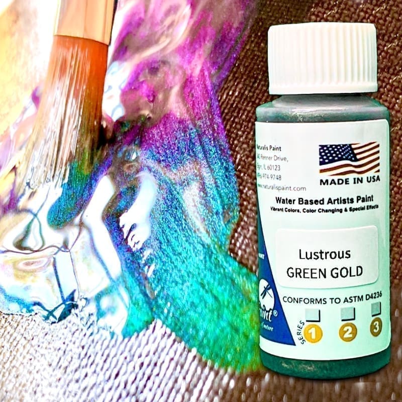 Close-up of a paintbrush blending green and purple paint beside a bottle of Lustrous Gold Green artist paint. The label mentions "water-based artists paint" and "made in USA.