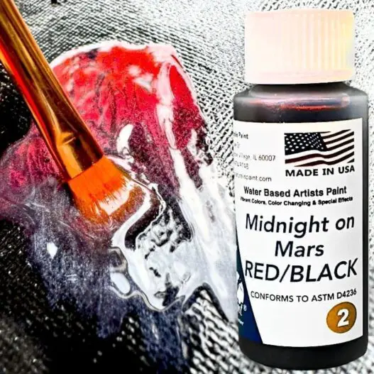 Close-up of a paintbrush applying red and black water-based paint from a labeled bottle called "Midnight on Mars Red Black" on a textured surface. The paint bottle features an American flag and manufacturing details.