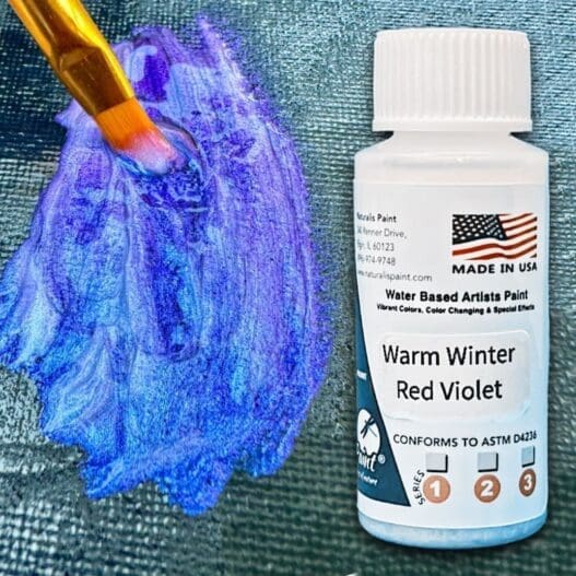 A paintbrush applies blue-violet paint onto a surface. Next to it is a bottle labeled "Warm Winter Red Violet," a water-based artist paint made in the USA by Warm Winter.