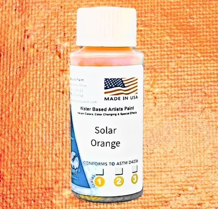 A small bottle of Solar Orange water-based artist's paint with a white cap and label showcasing an American flag, product details, and ASTM certification icons on an orange textured background.